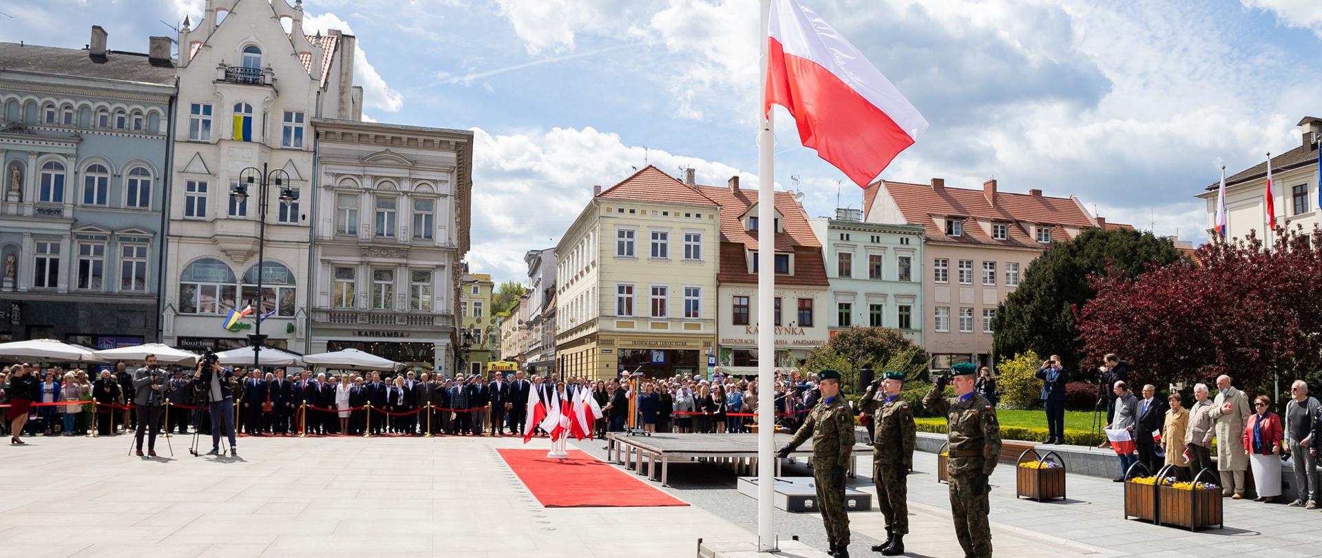 Obchody na płycie Starego Rynku w Bydgoszczy. Z lewej strony trzech żołnierzy w zielonych mundurach stojących przy maszcie flagowym, na którym w połowie wysokości powiewa flaga państwowa. Z lewej w tle widać zgromadzonych na uroczystościach ludzi stojących przy czerwonej taśmie. W tle pierzeje kamienic Starego Rynku w Bydgoszczy. 