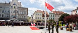 Obchody na płycie Starego Rynku w Bydgoszczy. Z lewej strony trzech żołnierzy w zielonych mundurach stojących przy maszcie flagowym, na którym w połowie wysokości powiewa flaga państwowa. Z lewej w tle widać zgromadzonych na uroczystościach ludzi stojących przy czerwonej taśmie. W tle pierzeje kamienic Starego Rynku w Bydgoszczy. 