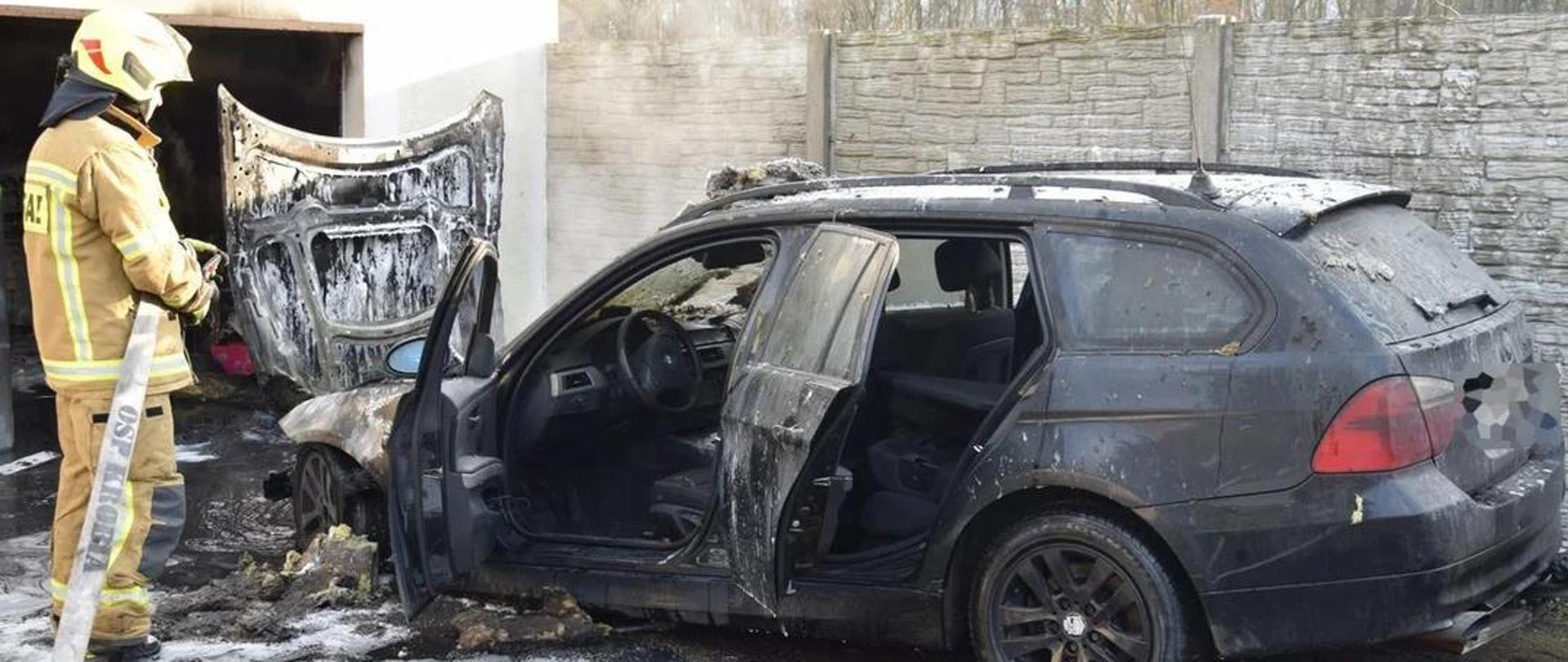 Zdjęcie przedstawia spalony samochód marki bmw oraz druha OSP trzymającego w rękach prądownice. 