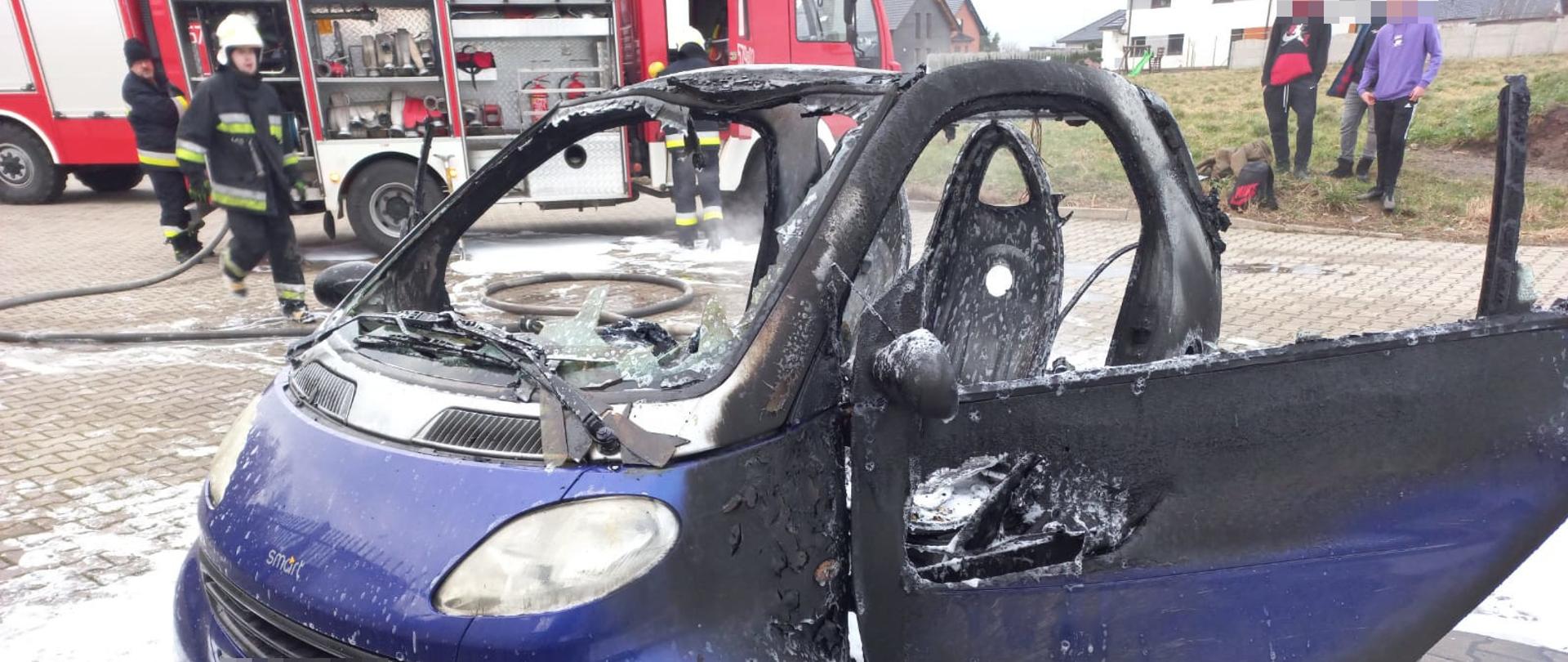 Pożar smarta. Parking przy Orliku w Bojanowie. Na pierwszym planie spalony pojazd. W tle wóz strażacki, strażacy i osoby postronne oraz budynki. Wokół samochodu pozostałości piany gaśniczej. 