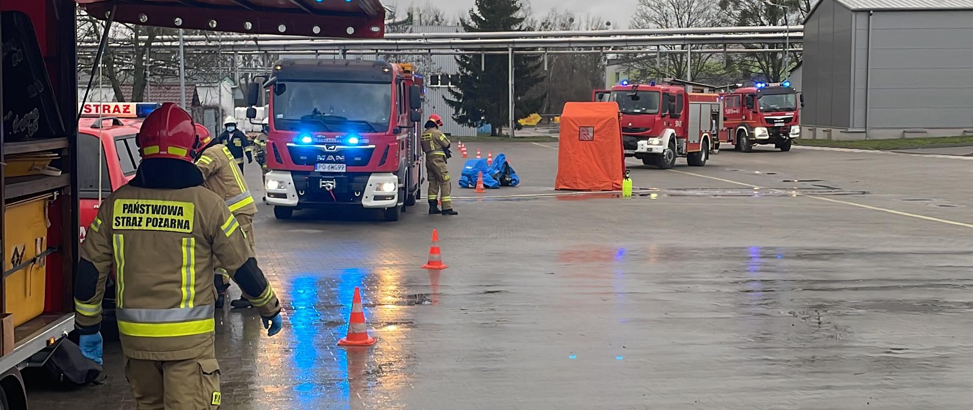 Przygotowanie do wejścia do działań podstawowych przez strażaków. na zdjęciu widać ustawione samochody pożarnicze kolor czerwonego wygrodzenie tereny(strefy) pachołkami drogowymi. Na pierwszym planie strażak stoi pod zadaszenie z czerwonego koloru po lewej stronie ma wyposażenie przyczepki chemicznej