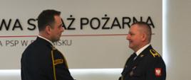 Mężczyźni w umundurowaniu wyjściowym. Komendant ściska rękę emeryta (strażaka).