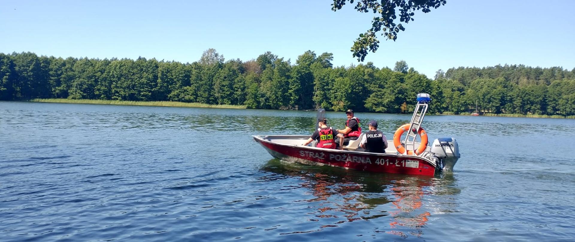 Zdjęcie przedstawia łódkę strażacką na jeziorze. W łódce znajduje się patrol składający się z strażaków i policjanta. Niebo jest bezchmurne. Na linii jeziora ciągnie się las