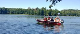 Zdjęcie przedstawia łódkę strażacką na jeziorze. W łódce znajduje się patrol składający się z strażaków i policjanta. Niebo jest bezchmurne. Na linii jeziora ciągnie się las