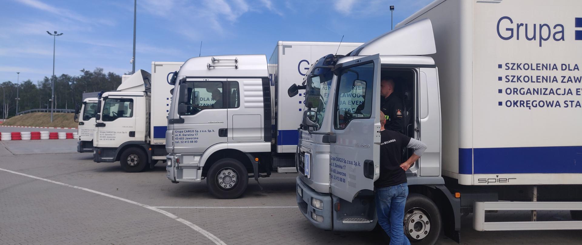 Na placu wyłożonym kostką stoją obok siebie cztery białe ciężarówki z naczepami. W kabinach siedzą kierowcy, obok pierwszego auta przy otwartych drzwiach stoi instruktor. 