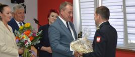 Świetlica komendy. Przedstawiciele władz samorządowych składają podziękowania i życzenia komendantowi powiatowemu. Burmistrz Jutrosina wręcza komendantowi prezent. Za nim z bukietami kwiatów w rękach stoją inni samorządowcy.