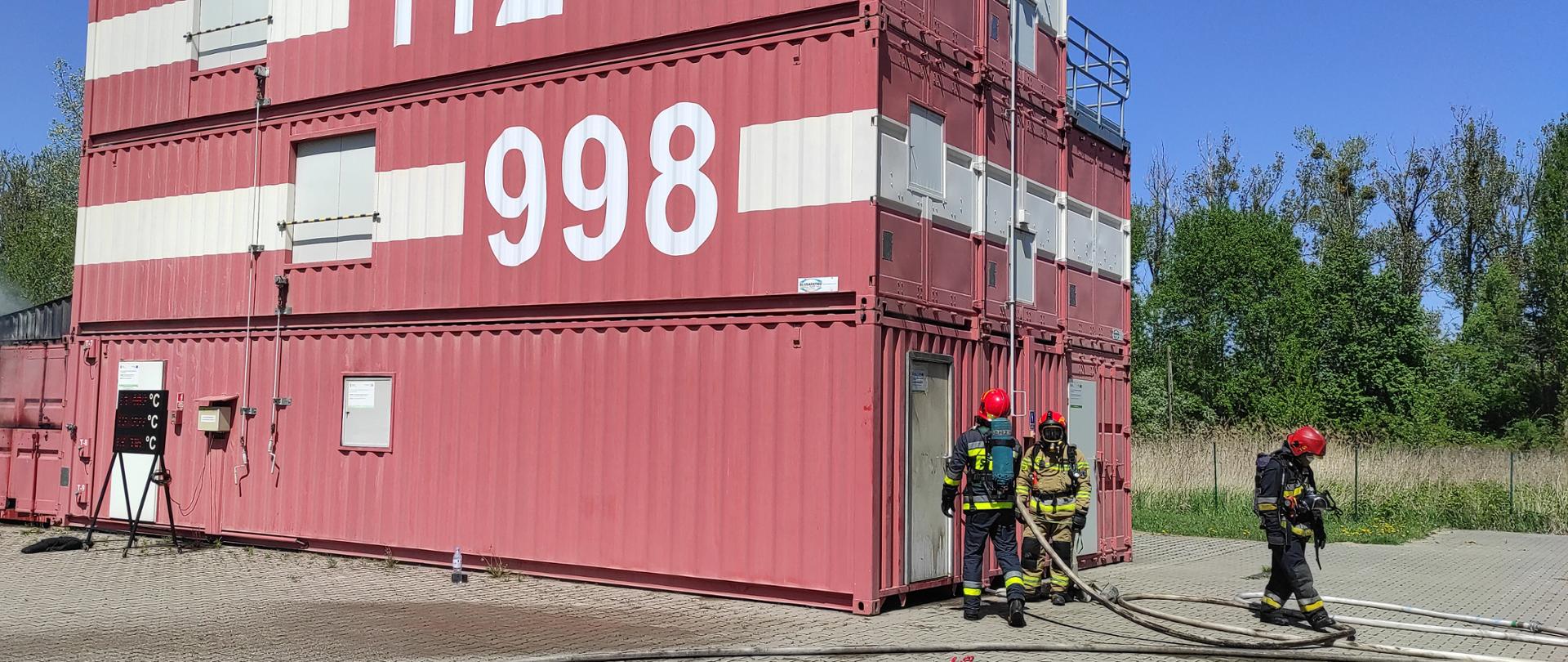 Na pierwszym planie widać ćwiczących strażaków ubranych w ubrania specjalne koloru piaskowego lub granatowego z odblaskowymi elementami oraz w czerwonych hełmach na głowie oraz założonymi maskami. Na plecach strażacy mają aparaty powietrzne butlowe. Widać strażaków ćwiczących przed czerwonobiałym trenażerem kontenerowym z którego wydobywa się jasnobrązowy dym. Na podłożu trawiastym i wybrukowanym widać rozwinięte węże strażackie i armaturę.