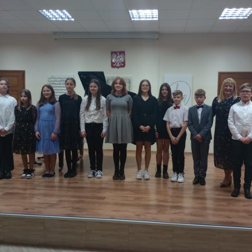Zdjęcie zbiorowe przedstawiające uczestników koncertu klasy fortepianu dodatkowego. Na brązowej drewnianej scenie stoją uczniowie i uczennice których jest 11, z prawej strony wśród nich stoi ich nauczycielka. W tle widać klapę fortepianu oraz wiszące godło państwowe.