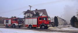 Samochód strażacki stoi przed zabudowaniami gospodarczymi, w których doszło do pożaru. Dookoła pozostałe zabudowania. Zdjęcie wykonane w porze zimowej.