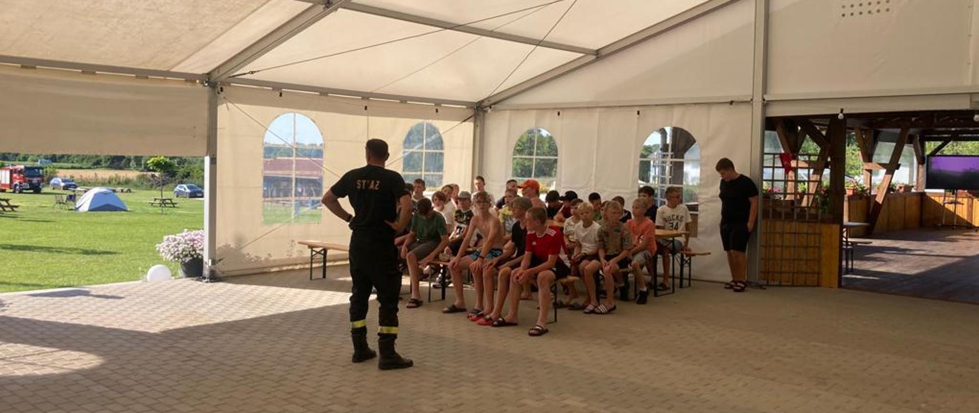 Na zdjęciu strażak przed grupą dzieci biorących udział w turnusie obozowym
