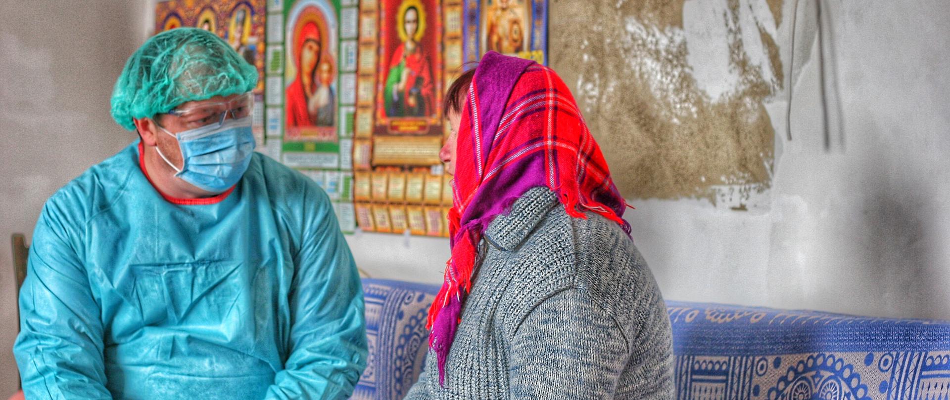 Lekarza w stroju ochronnym, maseczce i goglach rozmawia z pacjentką w kolorowej chuście w domu, w którym widnieją obrazy świętych.