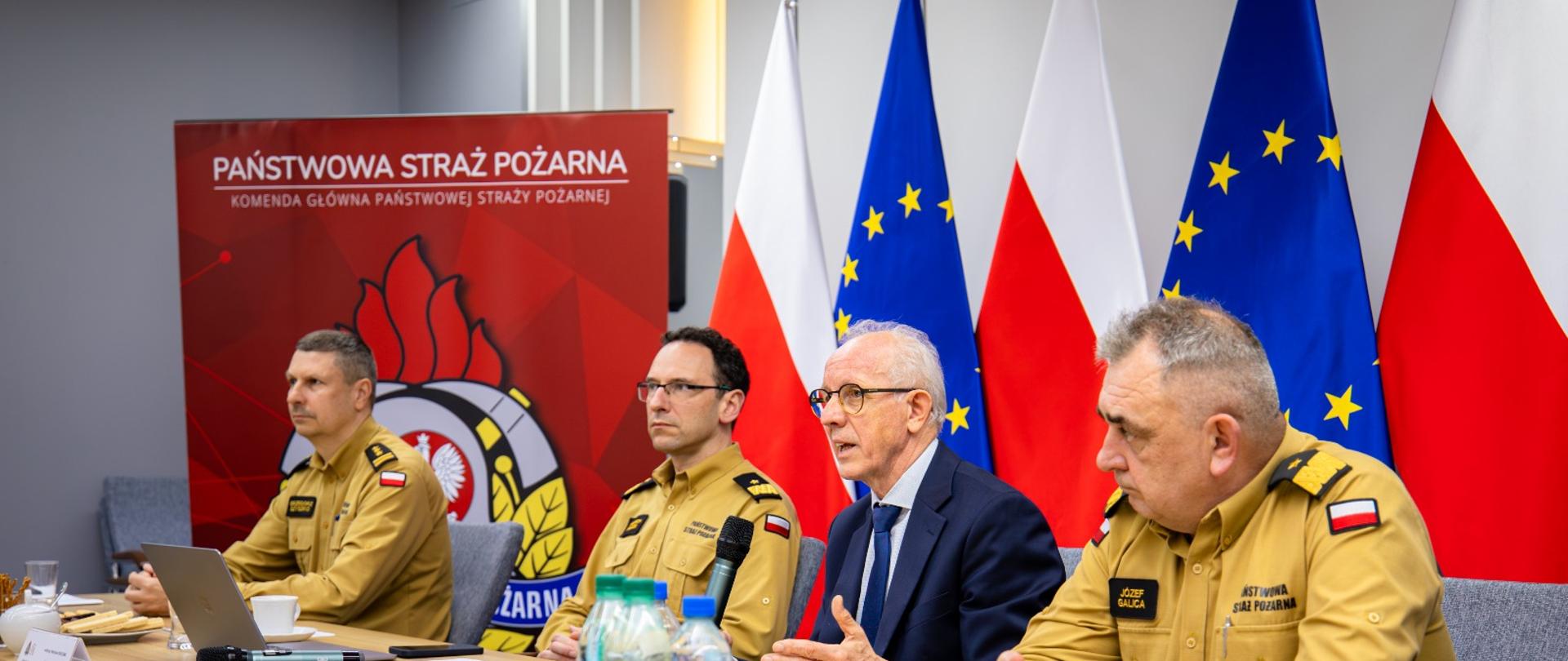 Szeroki kadr sali konferencyjnej, w której starszy mężczyzna w garniturze mówi do mikrofonu, otoczony przez wysokich rangą oficerów PSP w mundurach piaskowych. Na stole znajdują się butelki z wodą, dokumenty i notatniki. W tle widoczne są flagi oraz logo PSP.