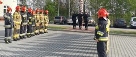 Na placu przed Jednostka Ratowniczo Gaśnicza nr 2 stoją strażacy w dwóch szpalerach naprzeciw siebie na uroczystej zbiórce. Pomiędzy szpalerami stoi dowódca. Strażacy ubrani są w stroje bojowe. Za szpalerami widoczny jest maszt na który poczet flagowy zawiesza biało-czerwona flagę.