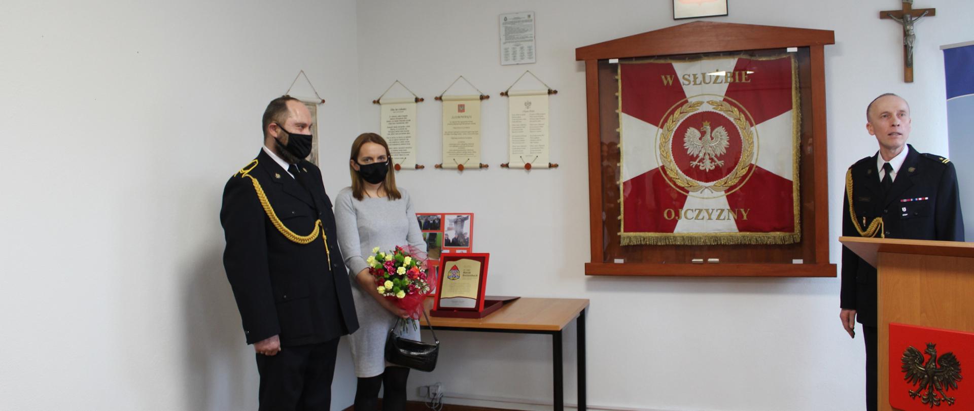 Zdjęcie przedstawia 2 mężczyzn ubranych w czarne garnitury z ozdobnym złotym sznurem oraz jedna kobieta ubrana na szaro. Obok niej znajduję się stół z prezentami, sztandar KP PSP Świdwin, mównica, Godło rzeczypospolitej Polskiej oraz Krzyż.