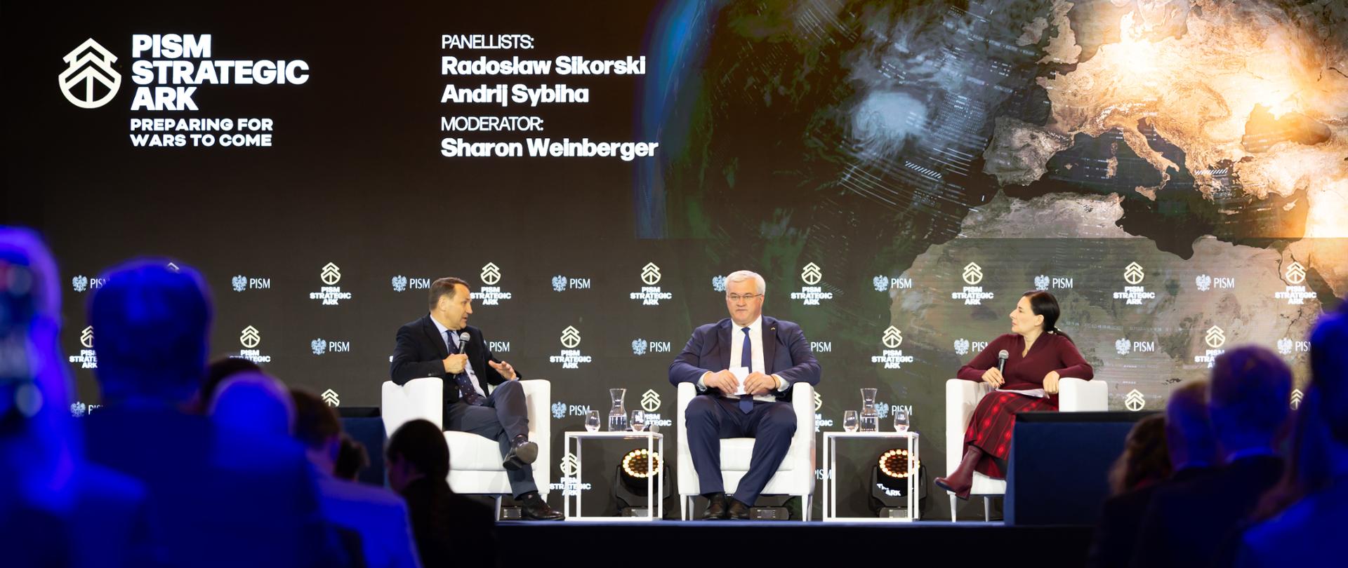 The photo shows three people—two men and a woman. They are sitting on white chairs on stage. They are speaking into microphones. Behind them, in the background, is the sign “PISM Strategic Ark.”