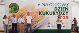 Grupa osób stoi na scenie podczas V Narodowego Dnia Kukurydzy 2025 w Smolicach, przed dużym banerem z wizerunkiem kolby kukurydzy i logotypami organizatorów. Jedna z osób przemawia do mikrofonu.