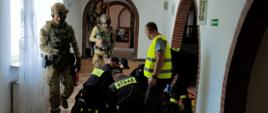 Na zdjęciu widać strażaków klęczących przed osobą poszkodowaną w pomieszczeniu. Obok nich stoją żołnierze z bronią. Obok strażaków stoi także instruktor w kamizelce. Zdjęcie wykonane w pomieszczeniu. 