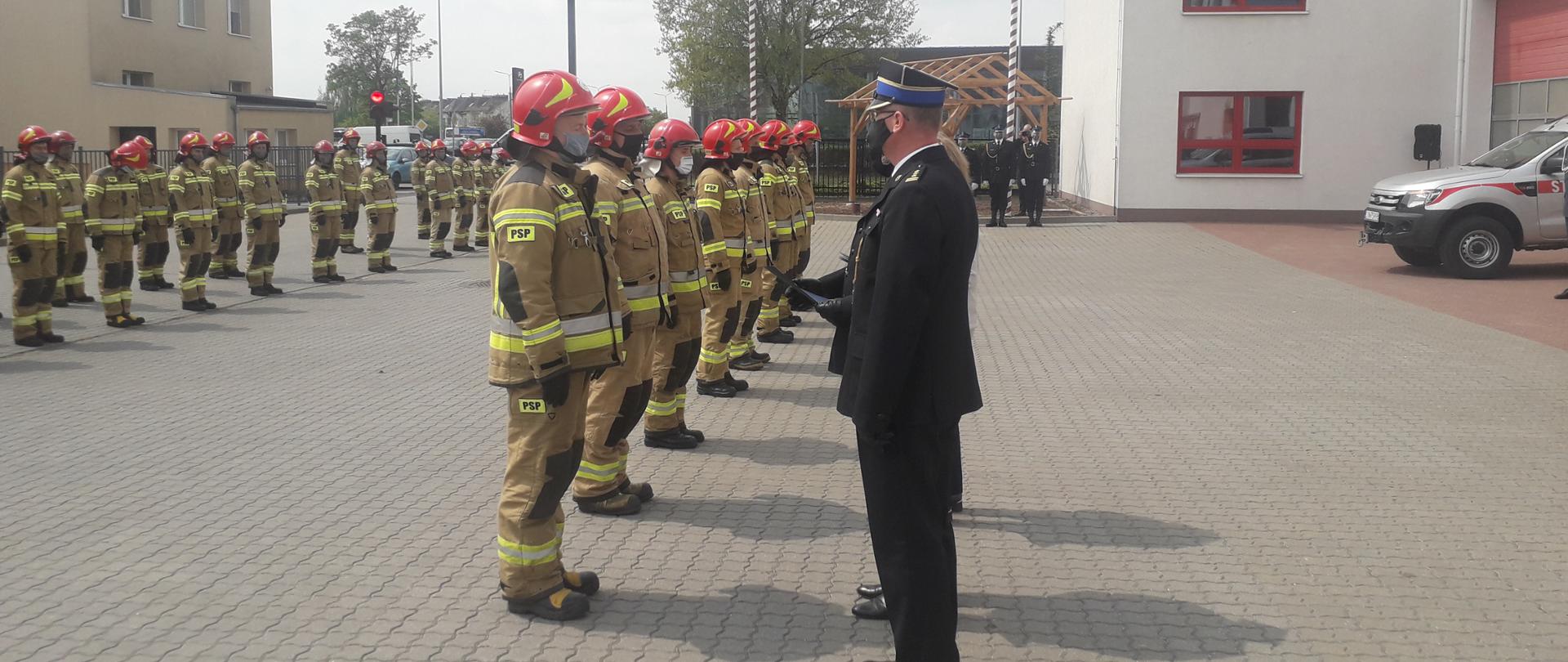 Na placu apelowym stoi pododdział strażaków w umundurowaniu bojowym. Na pierwszym planie komendant powiatowy wręcza w wyróżnienia. W tle poczet flagowy.