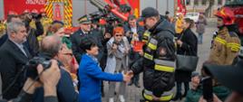 Pani poseł składa gratulacje strażakowi. Wokół zgromadzeni goście, w tym dziennikarze. W tle kilka pojazdów pożarniczych w ramach ekspozycji.