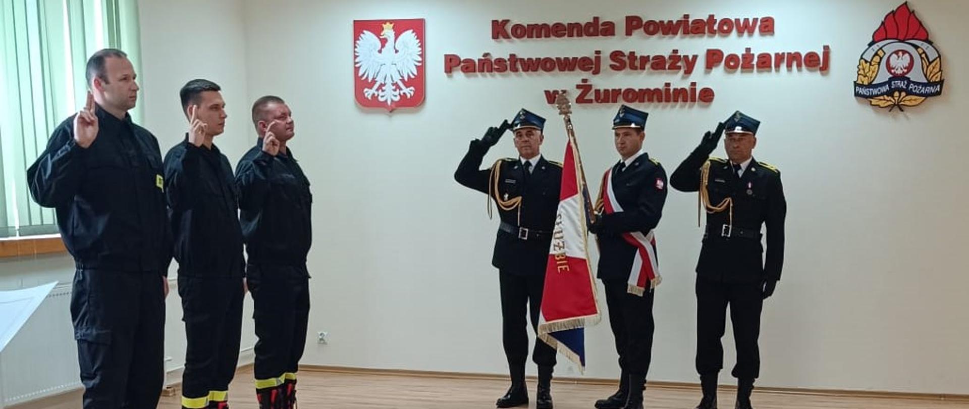 Na zdjęciu widzimy dalszy ciąg ceremonii ślubowania. Po lewej stronie znajdują się adepci, którzy wciąż mają podniesione dłonie w geście przysięgi. Na środku ściany widnieje napis "Komenda Powiatowa Państwowej Straży Pożarnej w Żurominie", co wskazuje na miejsce, w którym odbywa się ceremonia. Po obu stronach napisu widoczne są godła - polskie godło państwowe oraz godło straży pożarnej. Całe tło podkreśla formalny i uroczysty charakter wydarzenia.
