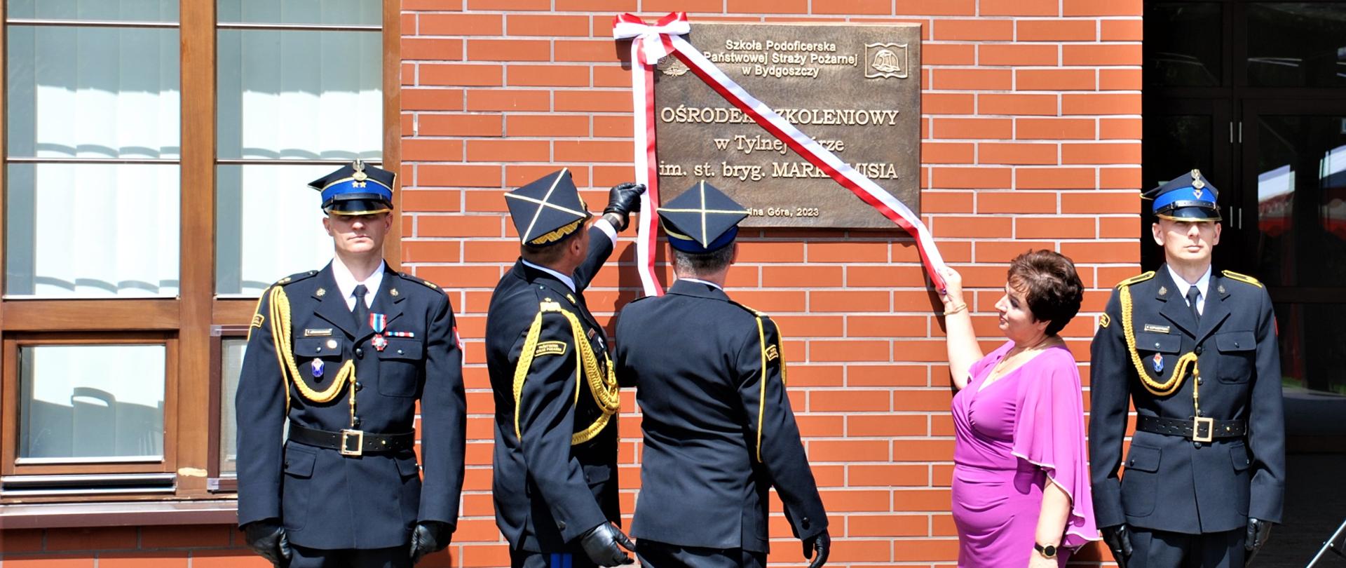 Zastępca komendanta głównego PSP wraz z komendantem szkoły i żoną śp. Marka Misia zawieszają czerwono - białą kokardę ze wstęgą na tablicy pamiątkowej po lewej i prawej stronie stoją strażacy w mundurach galowych 