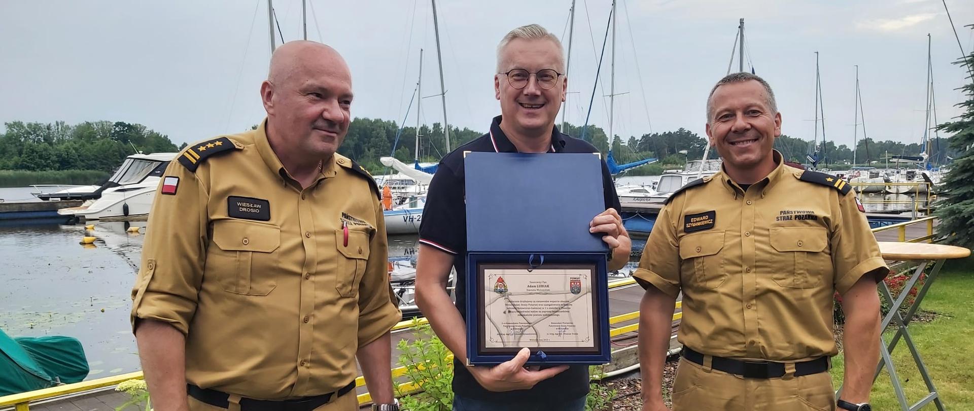Zdjęcie przedstawia Panów Komendantów wręczających ryngraf Panu Staroście. Strażacy ubrani są w umundurowanie służbowe. W tle zdjęcia widać zacumowane łodzie w porcie.