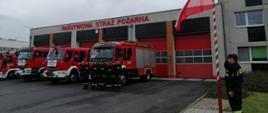 Strażacy w szregu stoją na tle pojazdów strażackich oraz budynku komendy.