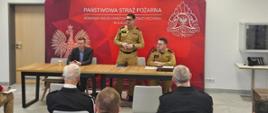Panowie Komendanci siedzą na krzesłach, na tle czerwonego baneru z napisem Państwowa Straż Pożarna i przemawiają do zgromadzonych gości.