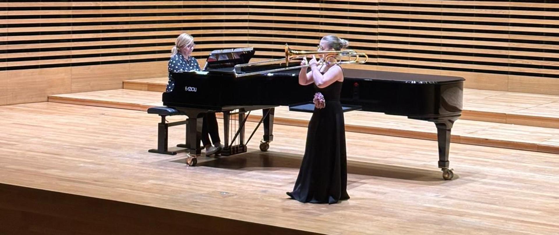 Dziewczyna gra na puzonie, kobieta akompaniuje na fortepianie.