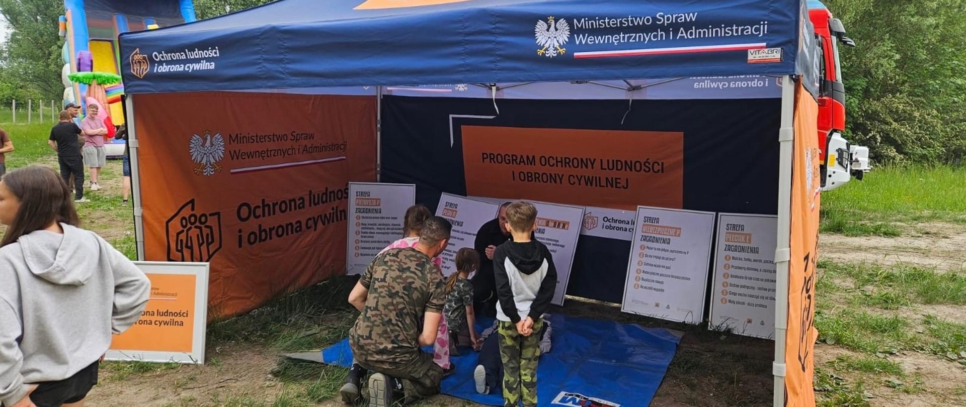 Widok z zewnątrz na namiot promocyjno-edukacyjny Ministerstwa Spraw Wewnętrznych i Administracji, poświęcony Ochronie Ludności i Obronie Cywilnej. Namiot ma ciemnoniebieski dach i pomarańczowe ścianki z orłem białym oraz hasłami edukacyjnymi. Wewnątrz namiotu, na niebieskiej macie, dorośli i dzieci uczestniczą w pokazie pierwszej pomocy. Po lewej stronie, tyłem do obiektywu, stoi dziewczyna w szarej bluzie, a w tle widać fragment czerwonego wozu strażackiego oraz dmuchaną zjeżdżalnię.