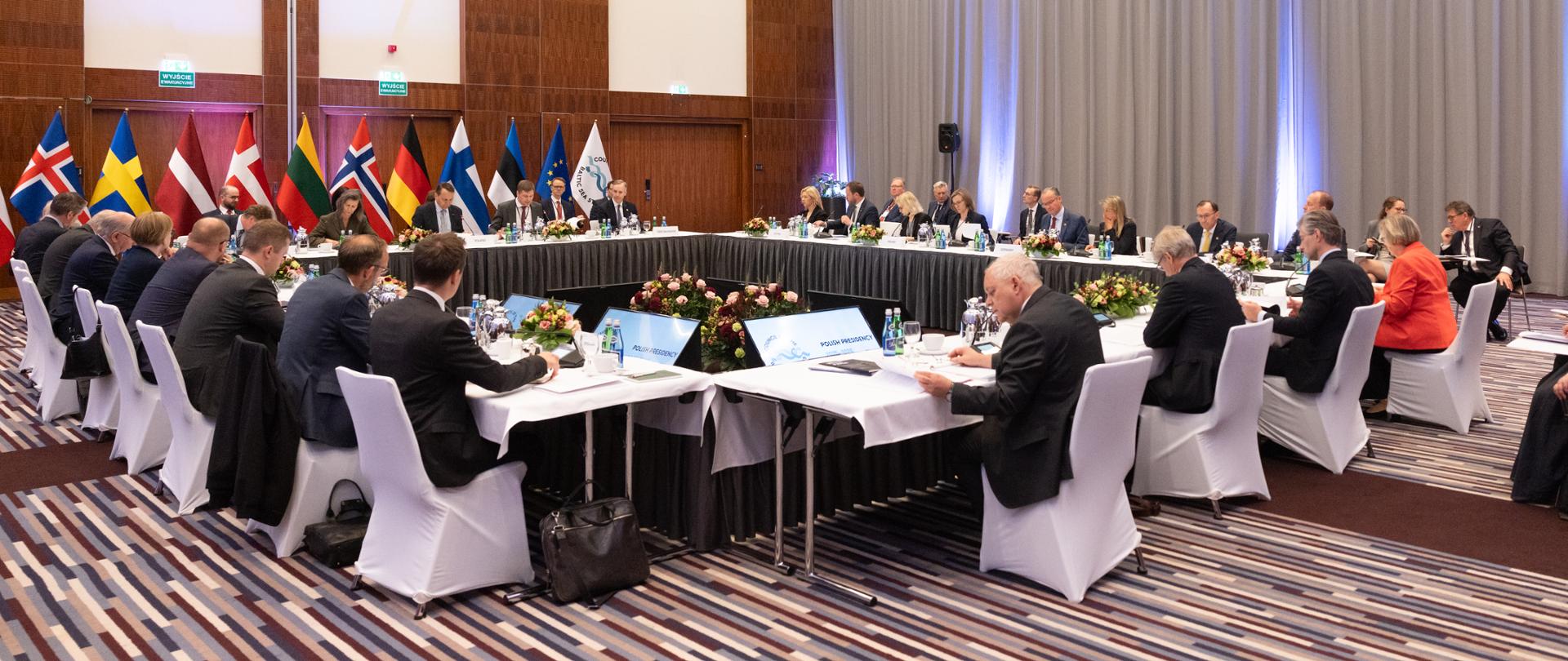 A group of people is sitting at tables arranged in a square. They have white chairs. The tables are covered with white tablecloths. There are flags in the background. 