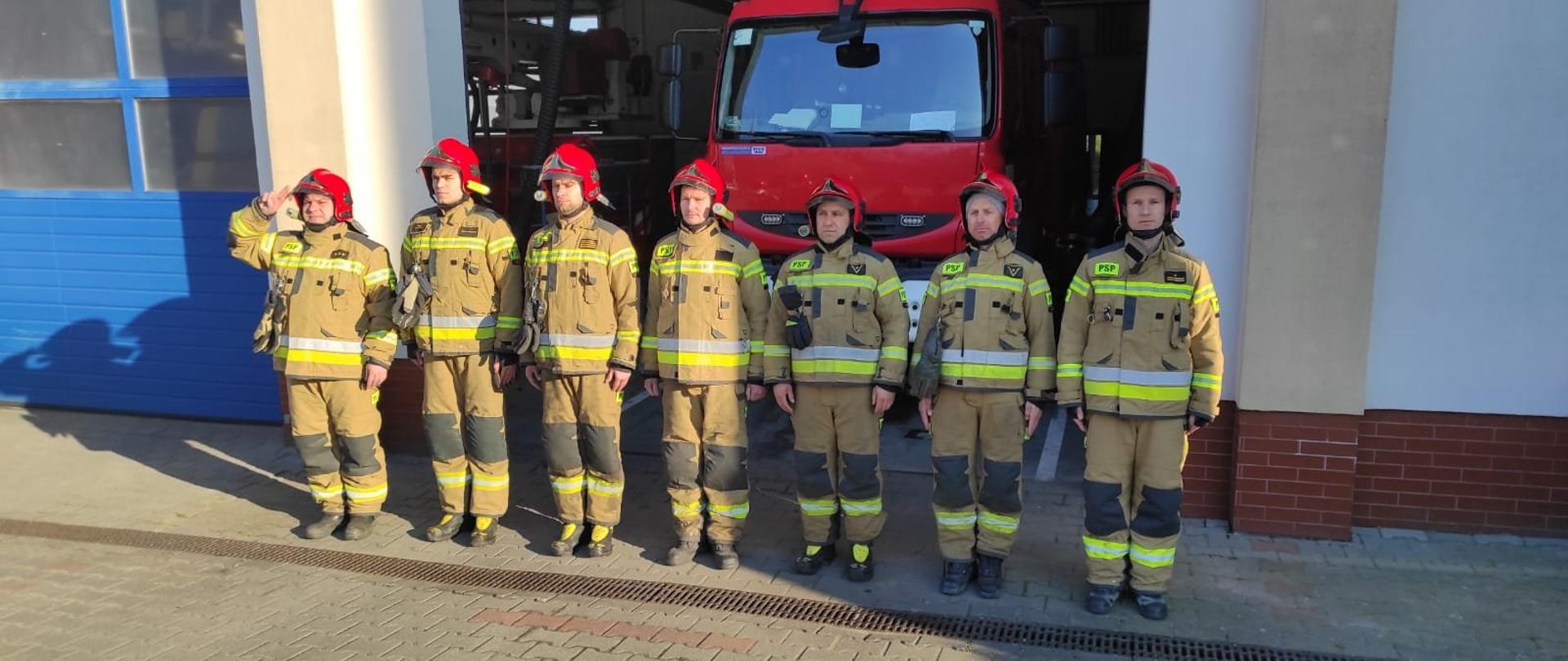 Na zdjęciu widoczny plac z polbruku i fragment budynku strażnicy. Przed otwartą bramą garażu siedmiu stojących w równym szeregu, wyprostowanych strażaków w piaskowych ubraniach bojowych i w czerwonych hełmach na głowie. Pierwszy od lewej salutuje. Za nimi widoczny przód strażackiego pojazdu.