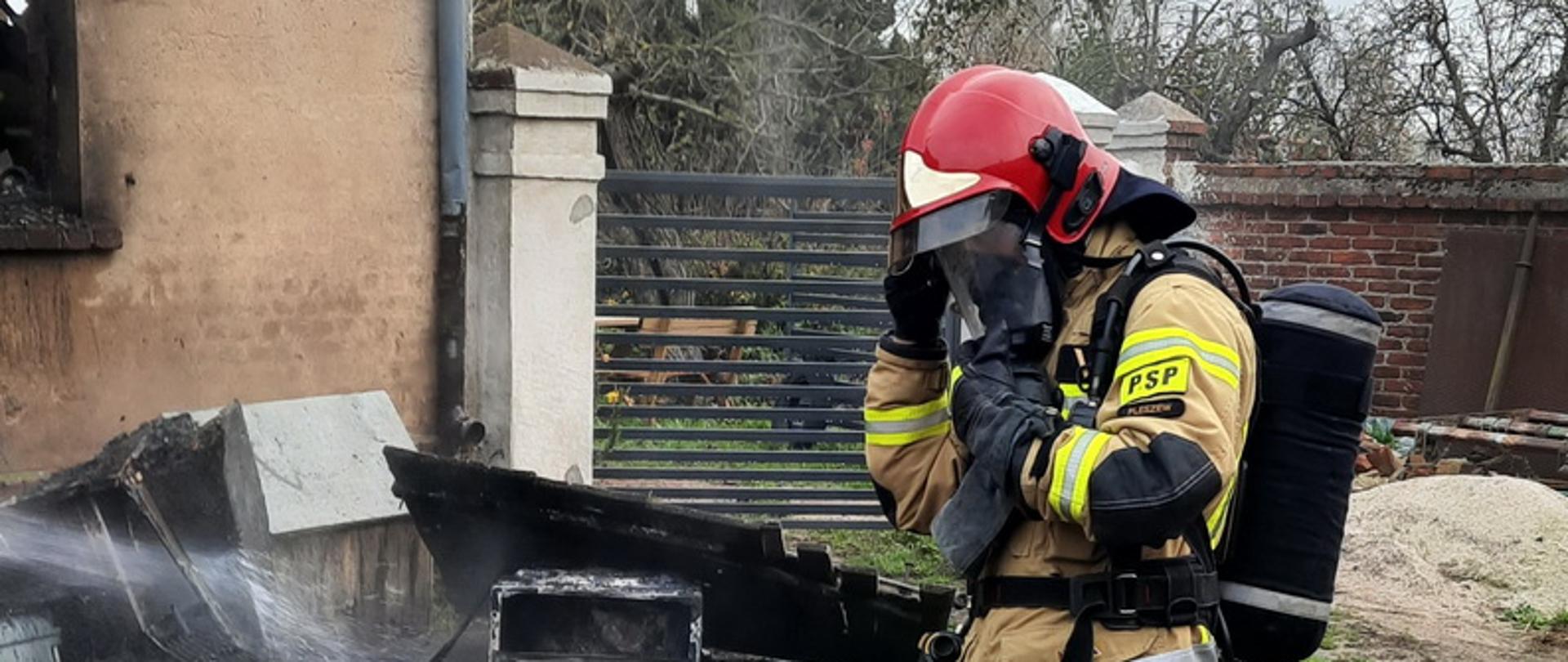 Pochylony strażak zdejmuje maskę aparatu ochrony dróg oddechowych, w tle widoczny spalony dom i leżące przed nim wyposażenie