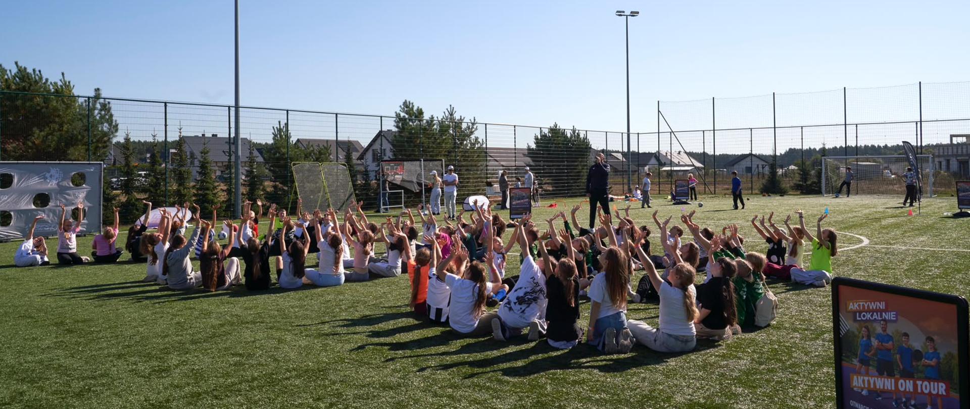 Zajęcia sportowe na Orliku. Dzieci siedzą na boisku w dwóch kółkach, patrzą w niebo i machają. W tle widać osoby uprawiające sporty. 