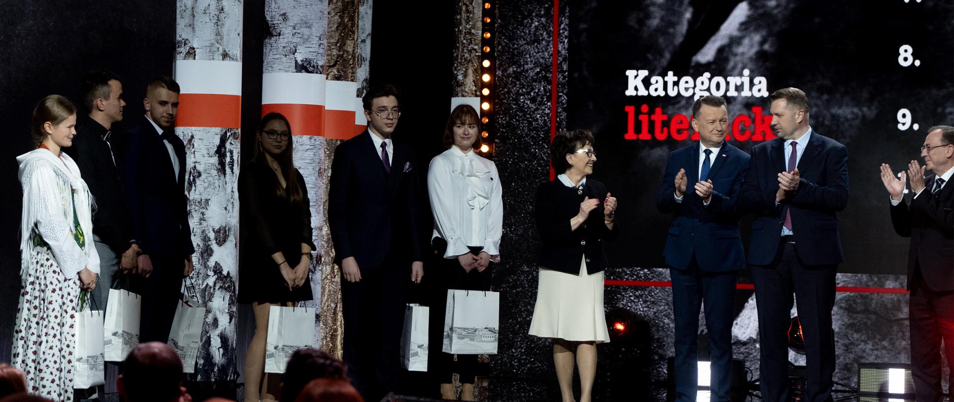 Na tle ściany w nieregularny wzór szarości i czerni stoi grupa elegancko ubranych osób, w rękach trzymają szare papierowe torby.