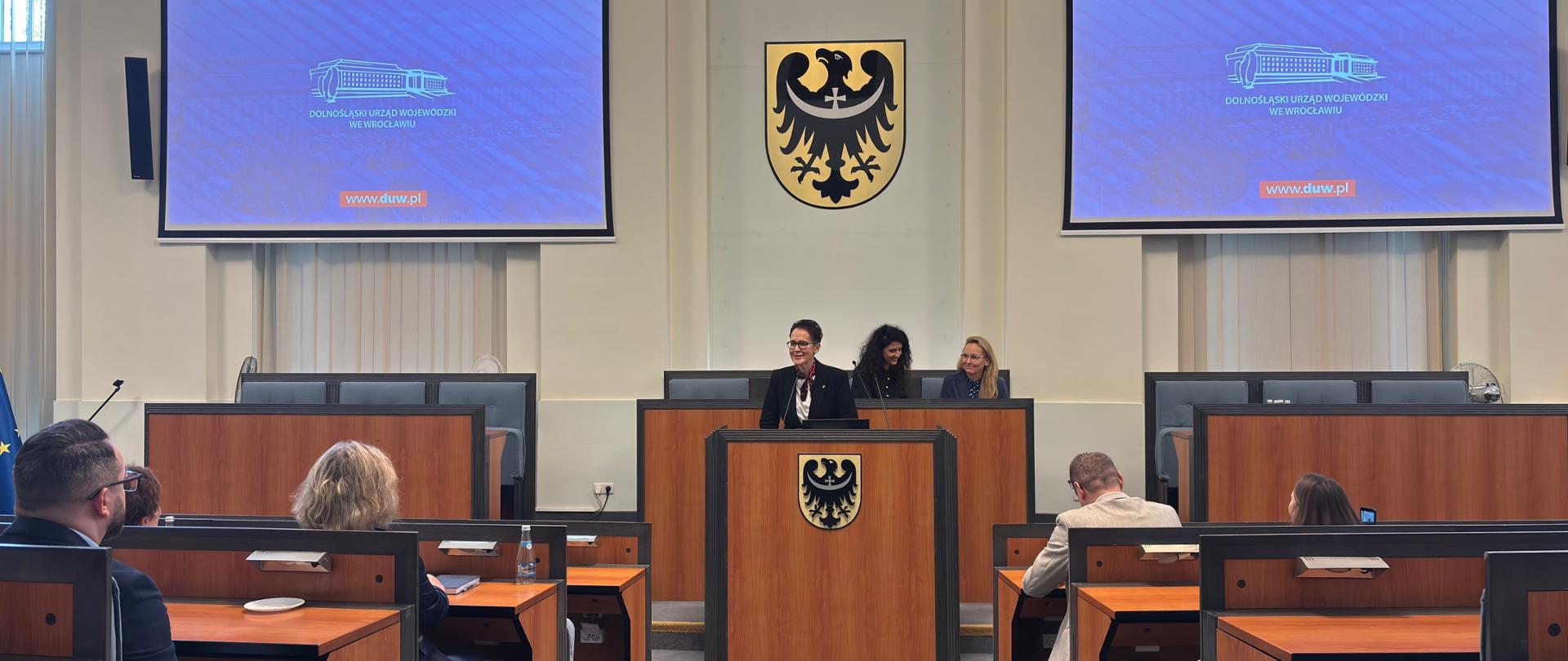 Szefowa służby cywilnej w przy mównicy, za nią herb Wrocławia - czarnego orła na żółtej tarczy
