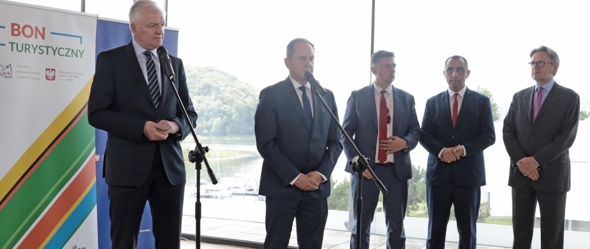 Wicepremier Jarosław Gowin, wiceminister Andrzej Gut-Mostowy stoją przy mikrofonach i mówią o Bonie Turystycznym. Obok przy szklanej ścianie stoi prezes POT Rafał Szlachta.