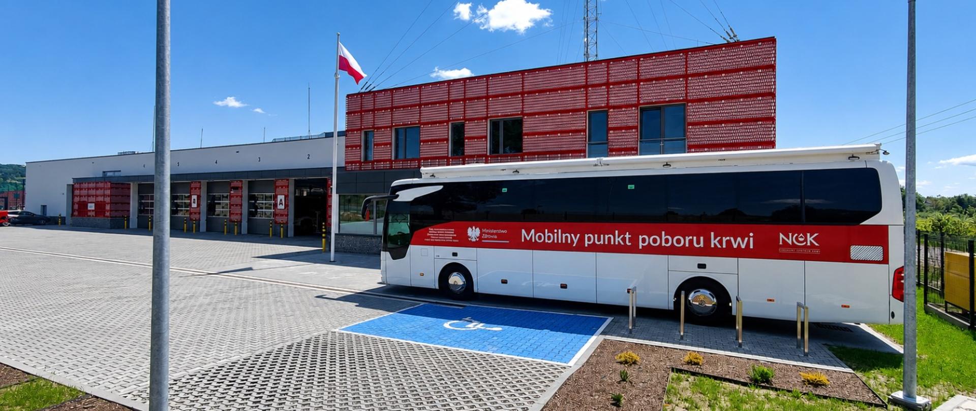Kolorowa fotografia wykonana w pogodny dzień obok budynku Komendy Powiatowej PSP w Brzozowie. Przedstawia autobus-krwiobus Regionalnego Centrum Krwiodawstwa i Krwiolecznictwa w Rzeszowie ustawiony przy wejściu do komendy, Obok widoczny maszt z flagą.