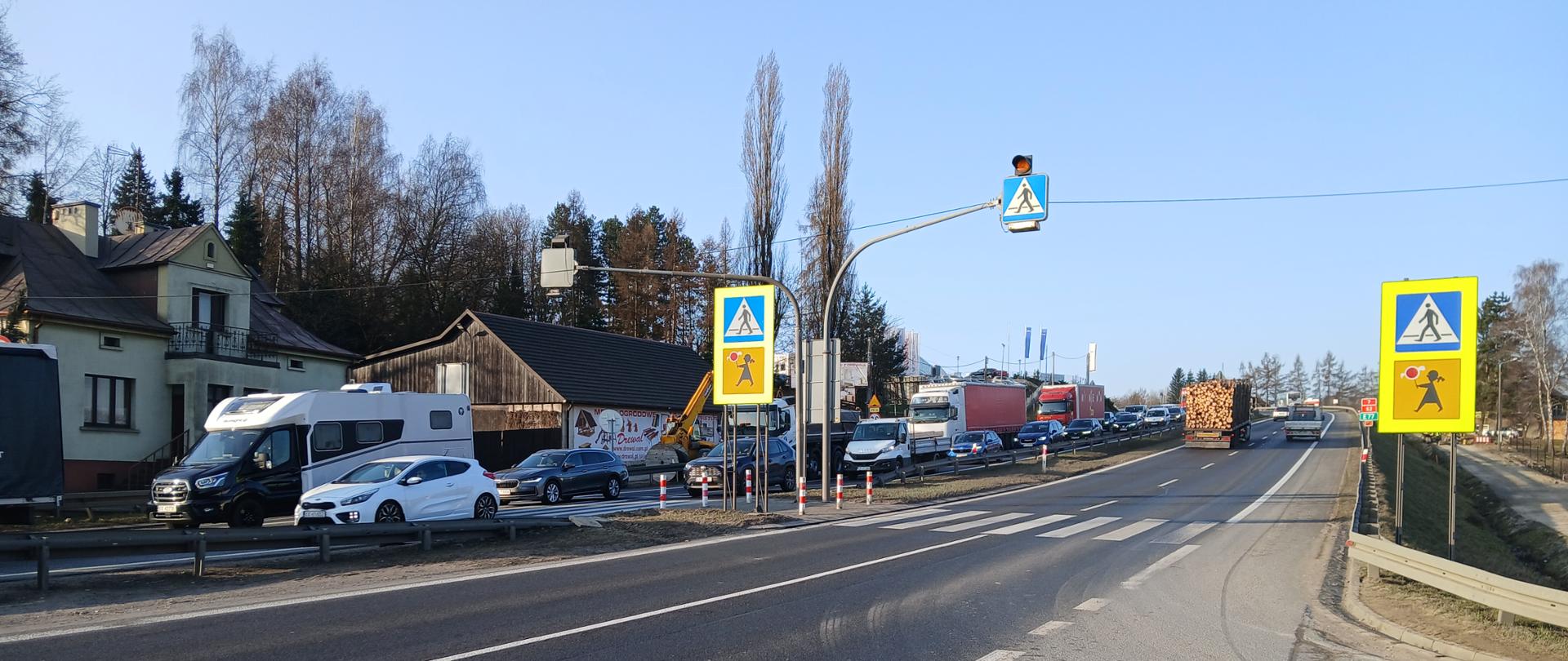 Przejście dla pieszych na dwujezdniowej drodze z wysepką dzielącą, wyposażone w sygnalizację świetlną z ostrzegawczymi sygnałami żółtymi. Widoczne oznakowanie pionowe przejścia oraz logotyp „Aktywny znak”. Na jezdni ruch samochodowy, w tym samochody osobowe i ciężarowe; po lewej stronie budynki mieszkalne i gospodarcze, po prawej pobocze z barierą ochronną. 