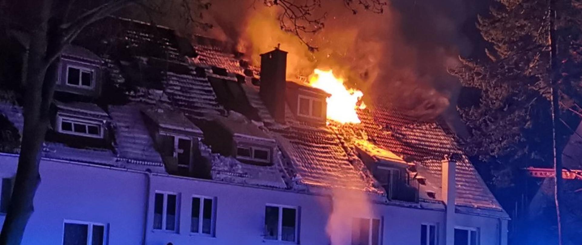 Dach budynku mieszkalnego, widoczne płomienie wydobywające się z jednego z okien.
