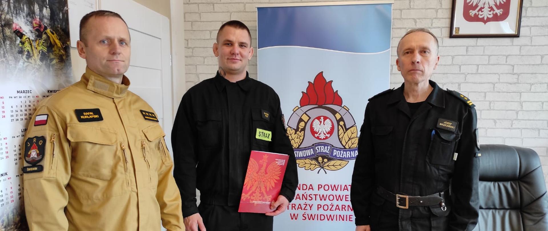 na zdjęciu po lewej ubrany w piaskowy strój koszarowy dowódca jednostki,na środku strażak ze świadectwem ukończenia kursu w dłoni, po prawej komendant jednostki ubrany na czarno 