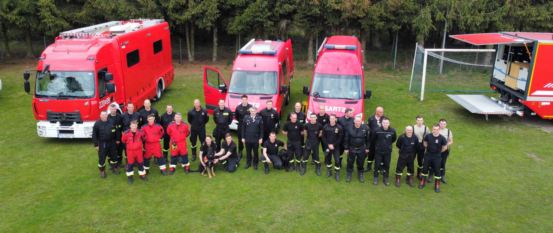 Zdjęcie przedstawia strażaków oraz sprzęt i pojazdy strażackie podczas pokazów Państwowej Straży Pożarnej.
W tle drzewa i płyta boiska sportowego.
