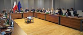 Przy ustawionym w czworokąt stole konferencyjnym siedzą uczestnicy spotkania i słuchają przemawiającego mężczyzny