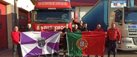 Dzień. W centralnej części zdjęcia dwa samochody ciężarowe ustawione na wprost. Kabina pierwszego w kolorze czerwonym, nad przednią szybą napis "Bombeiros" szarymi literami. Po prawej stronie stoi drugi TIR którego kabina jest w kolorze białym. Przed samochodami stoi grupka strażaków. Sześciu z nich to polscy strażacy ubrani w czarne mundury koszarowe. Pomiędzy nimi stoi czterech strażaków z Portugalii ubranych w czarne spodnie i czerwone bluzy. Strażacy trzymają dużą flagę Portugalii (prostokąt podzielonym na dwa pionowe pasy: zielony i czerwony. Na złączeniu pasów umieszczony jest herb Portugalii) oraz flagę miejscowości Anadia (prostokąt podzielony na biało fioletowe trójkąty, na środku umieszczony herb Anadi).