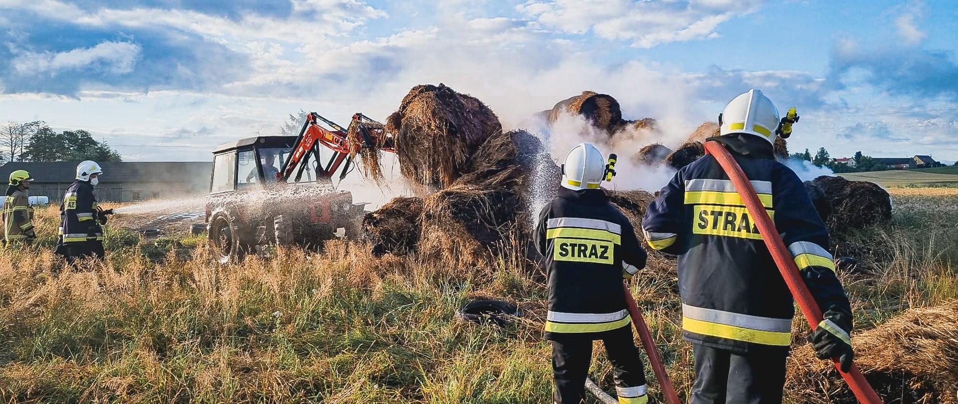 Strażacy w ubraniu specjalnym gaszą palący się stóg balotów słomy, z którego jest duże zadymienie. Na drugim planie traktor z ładowaczem czołowym, który przekłada nadpalone baloty.
