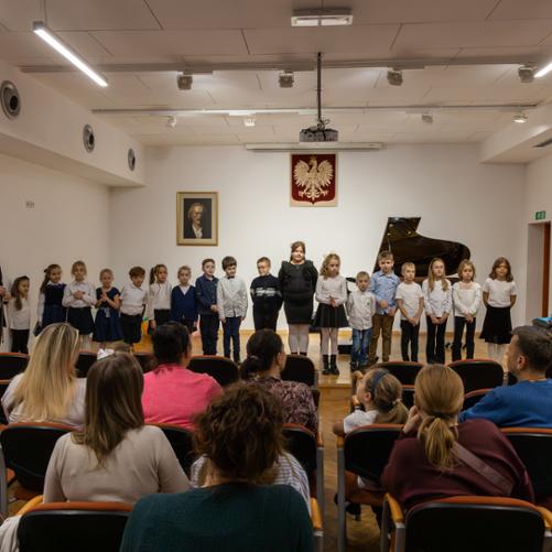 uczniowie klasy pierwszej wspólnie stojący na scenie w auli szkoły muzycznej w głubczycach, z lewej strony zdjęcia widać pana dyrektora szkoły, który pozuje wraz z uczniami, z prawej strony stoi pani przygotowująca uczniów do uroczystości pasowania, w dolnej części zdjęcia widać publiczność zgromadzoną na wydarzeniu