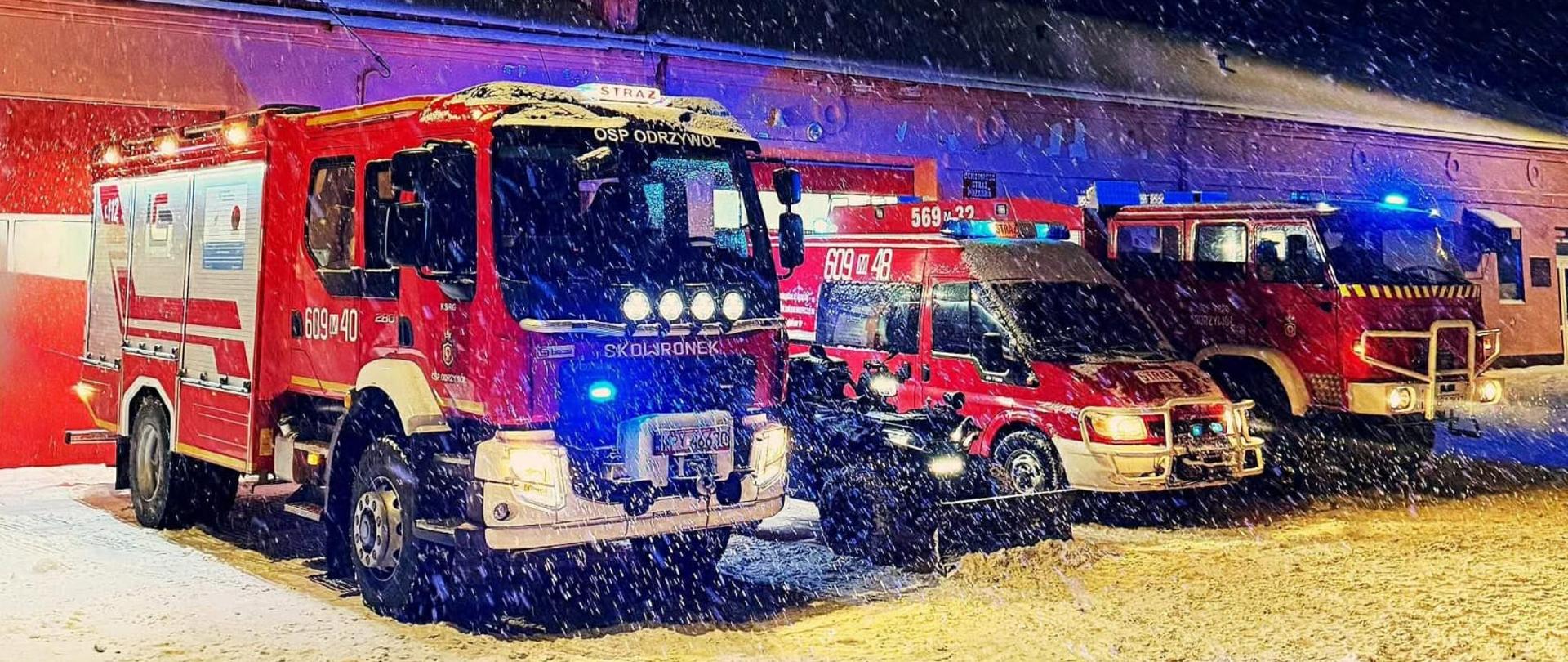 Zdjęcie przedstawia zimową, nocną scenerię przed remizą strażacką w miejscowości Odrzywół. Panują trudne warunki atmosferyczne – widoczny jest gęsty opad śniegu, który w świetle reflektorów tworzy efekt jasnych plamek na całym obrazie. Na pierwszym planie, na ośnieżonym placu, zaparkowane są pojazdy ratownicze Ochotniczej Straży Pożarnej (OSP) w Odrzywole:
Po lewej stronie: Nowoczesny, średni samochód ratowniczo-gaśniczy marki VOLVO (o numerach operacyjnych 609-40). Pojazd ma włączone światła mijania oraz intensywne, niebieskie sygnały błyskowe. Na masce widnieje napis „SKOWRONEK”.
W środku: Mniejszy wóz techniczny (prawdopodobnie marki Ford) oraz zaparkowany przed nim quad, również wyposażony w oświetlenie ostrzegawcze.
Po prawej stronie: Starszy typ wozu strażackiego - STAR 266, oświetlony niebieską łuną kogutów.
W tle widoczny jest parterowy budynek remizy z czerwonymi bramami garażowymi. Całość oświetlona jest mieszanką ciepłego światła lamp ulicznych oraz zimnego, niebieskiego blasku sygnałów alarmowych, co nadaje zdjęciu dynamiczny i surowy charakter.