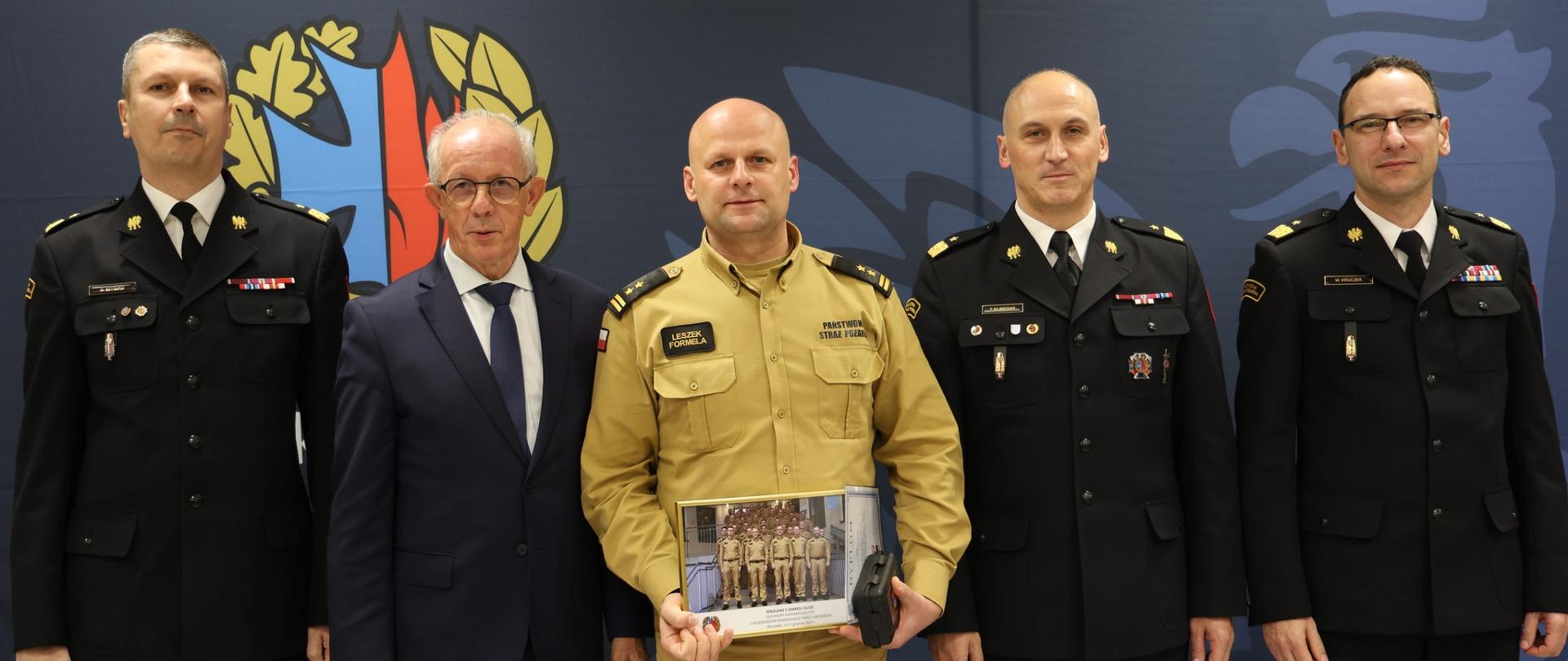 Brygadier Leszek Formela Komendant Powiatowy Państwowej Straży Pożarnej w Lęborku stoi na pamiątkowej ściance trzymając w dłoniach pamiątkowe zdjęcie uczestników szkolenia. Po lewej jego stronie stoi Pan Wiesław Leśniakiewicz – Podsekretarz Stanu w MSWiA i nadbryg. dr inż. Grzegorz Szyszko – Zastępca Komendanta Głównego PSP, po prawej jego stronie Pan nadbrygadier Tomasz Klimczak - Rektor Akademii Pożarniczej w Warszawie oraz nadbrygadier Wojciech Kruczek – Komendant Główny PSP.