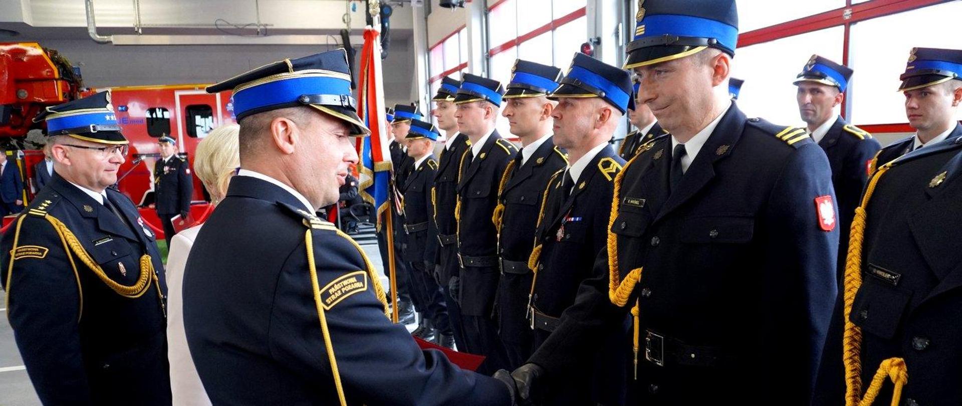 Zdjęcie zrobione wewnątrz garażu JRG, podczas uroczystych obchodów Dnia Strażaka. Oficer Państwowej Straży Pożarnej w stopniu starszego brygadiera gratuluje strażakowi wyróżnienia. Obok strażaka stoją uczestnicy uroczystości - strażacy Państwowej Straży Pożarnej - wszyscy ubrani są w mundury galowe. 