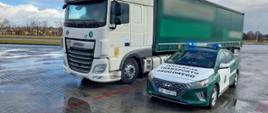 Od lewej: ciągnik siodłowy z podpiętą naczepą i stojący obok ciężarówki oznakowany radiowóz śląskiej Inspekcji Transportu Drogowego z włączonymi niebieskimi światłami uprzywilejowania w ruchu drogowym.